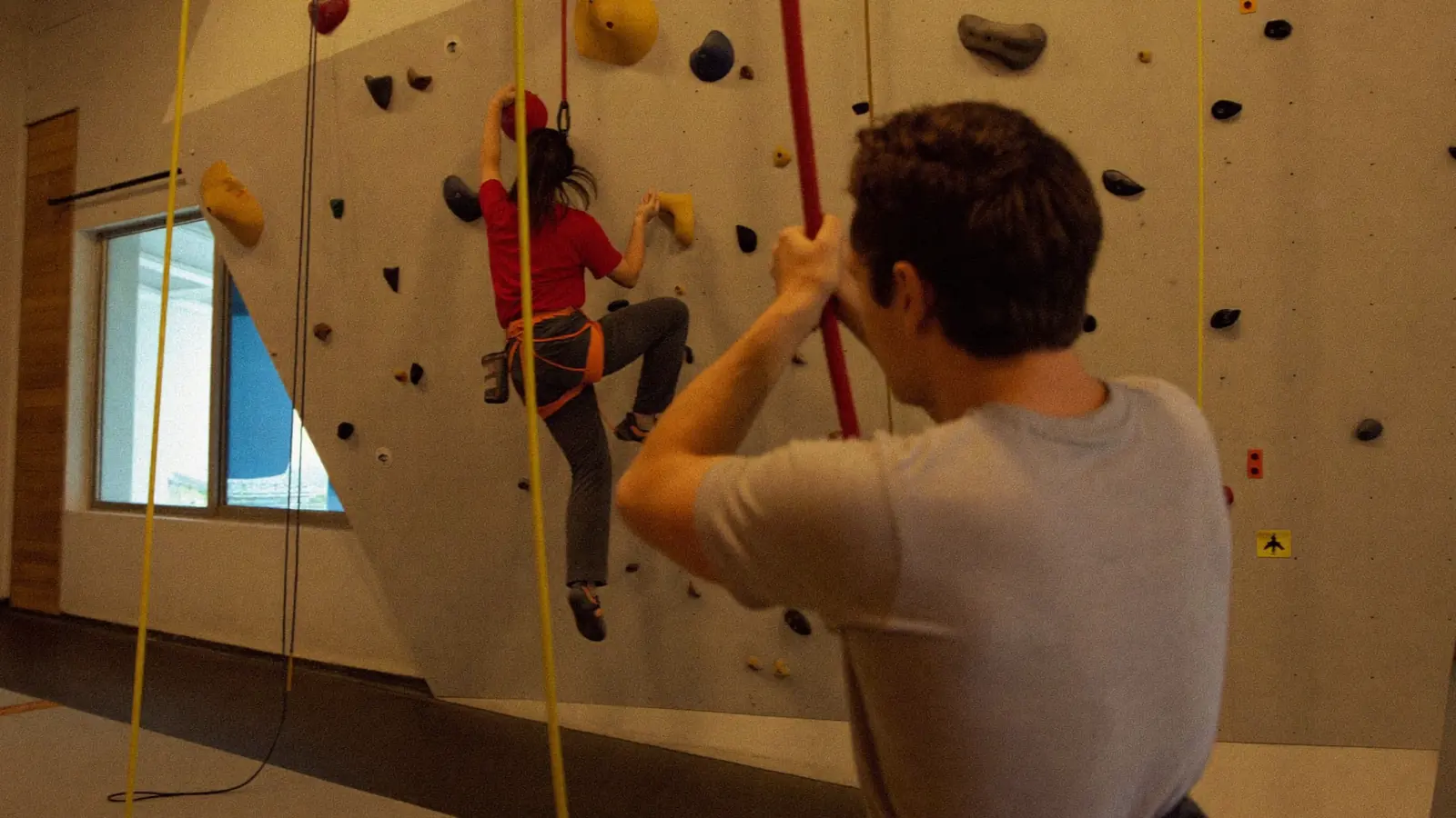 Istruttore di arrampicata che fa sicura a un adulto su una parete colorata in una palestra indoor, con una presa in primo piano