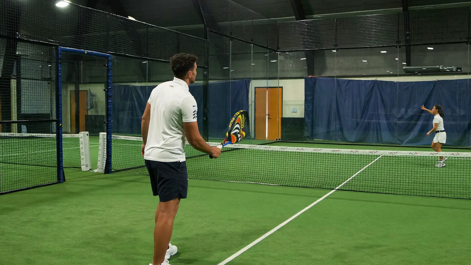 Istruttore che corregge il dritto di un adulto su un campo da padel indoor con pareti in vetro