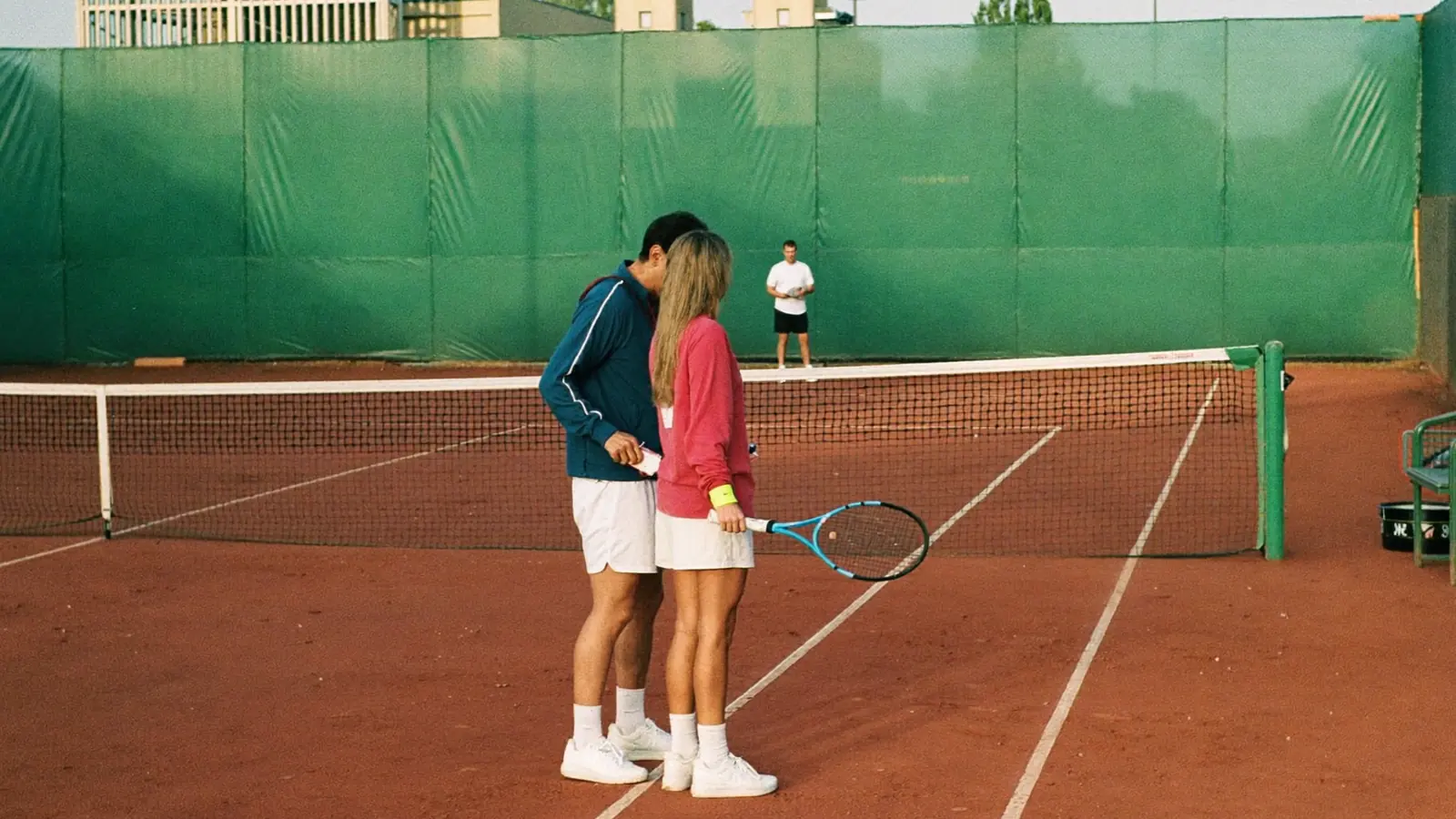 Istruttore di tennis che corregge il rovescio di un allievo adulto su un campo in terra rossa al mattino