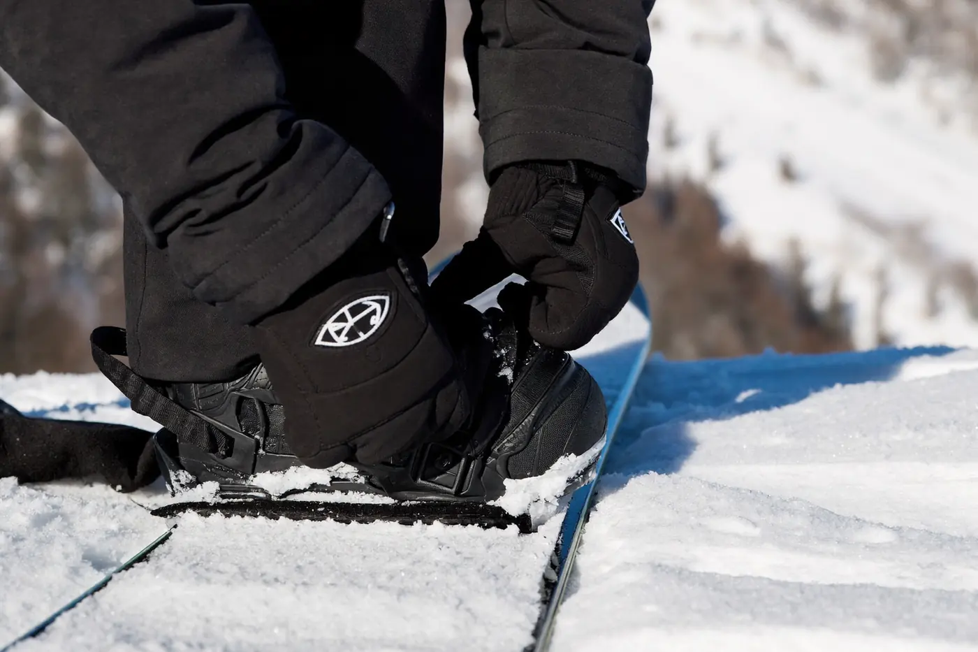 Dettaglio degli attacchi di una tavola da snowboard appoggiata sulla neve, con un gruppo sfocato sullo sfondo