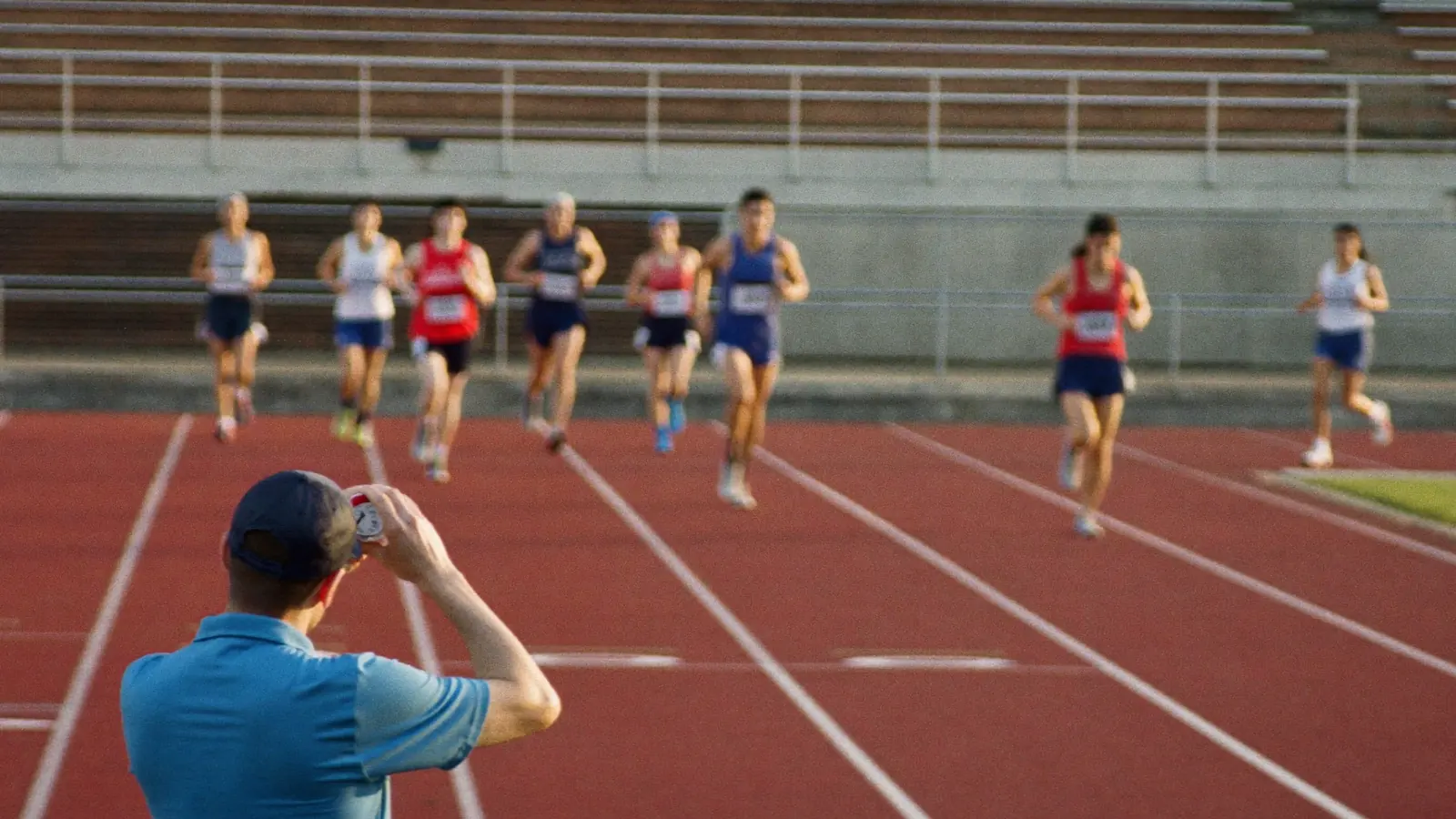 Tecnico di atletica leggera con cronometro in mano osserva un gruppo di atleti adulti sui blocchi di partenza di una pista rossa nel tardo pomeriggio