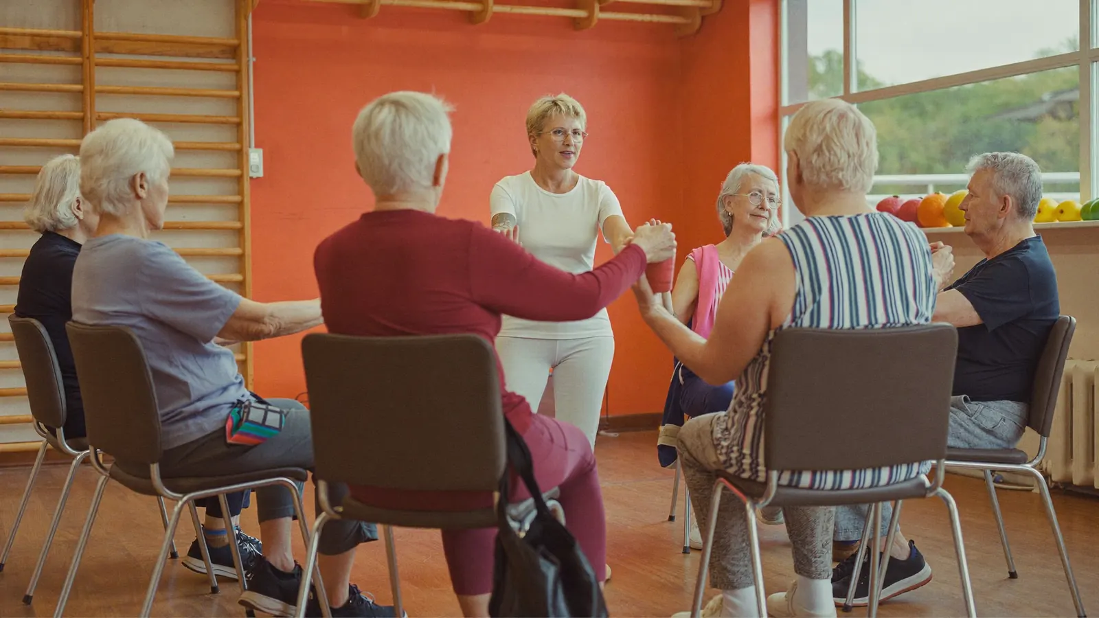 Gruppo di persone anziane sedute in cerchio su sedie in una sala luminosa, con l'istruttrice al centro che guida l'esercizio