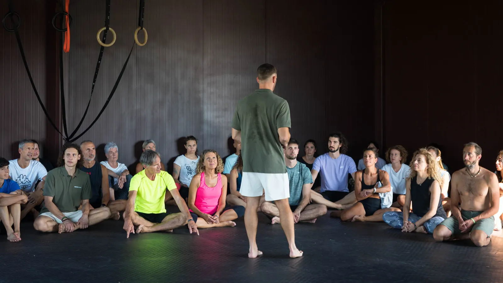 Coach in piedi che parla a un grande cerchio di persone sedute a terra in una sala con anelli da ginnastica appesi al soffitto