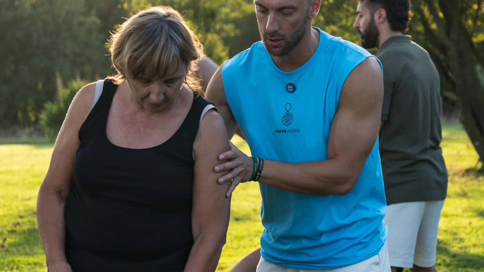 Coach in canotta azzurra che con la mano corregge la posizione della spalla di una donna matura, all'aperto su un prato