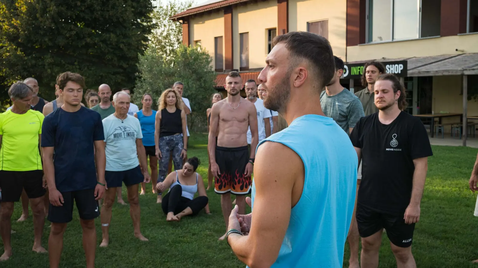 Coach in canotta azzurra parla con energia a un gruppo di persone in piedi all'aperto, davanti a un edificio in mattoni